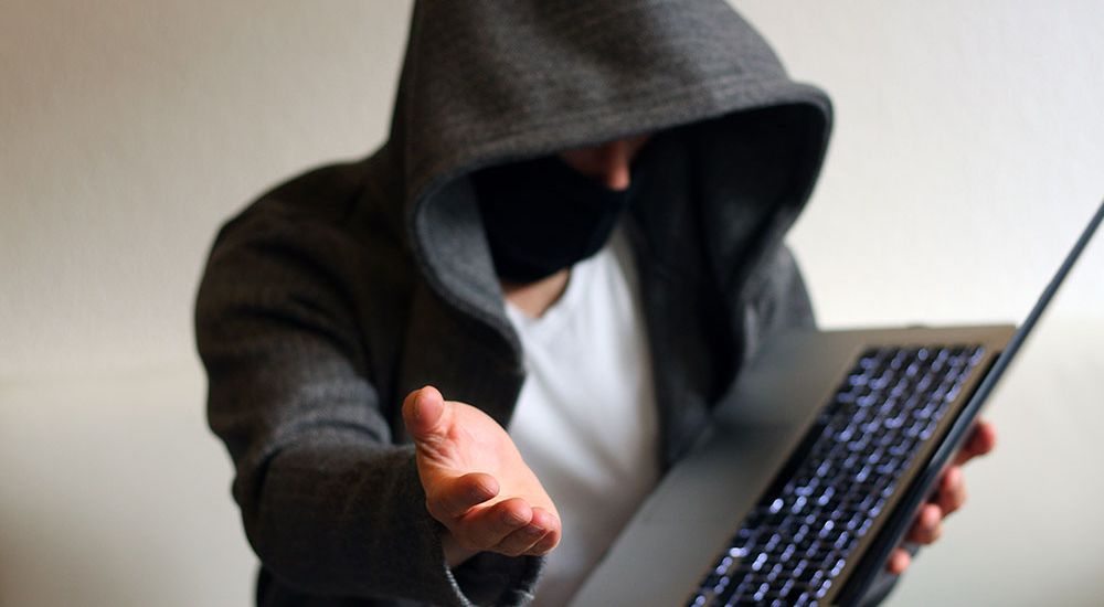 Hooded person with mask holding laptop, reaching out hand.