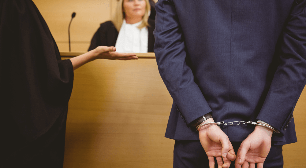 Defendant in handcuffs stands before judge in courtroom, legal proceeding in progress.