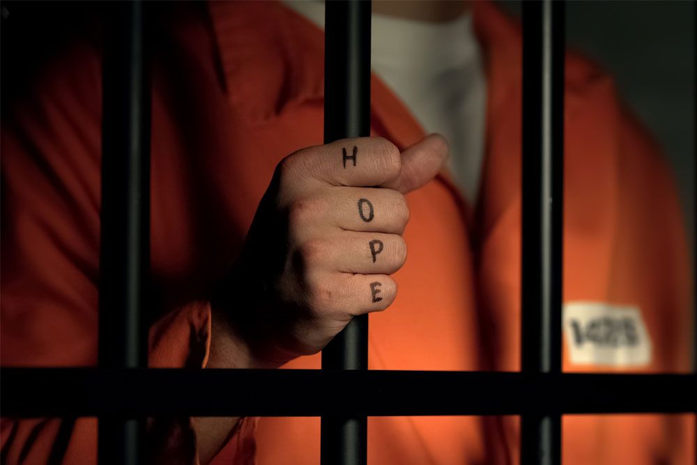 Inmate grips prison bars with Hope tattoo on fingers, wearing an orange jumpsuit.