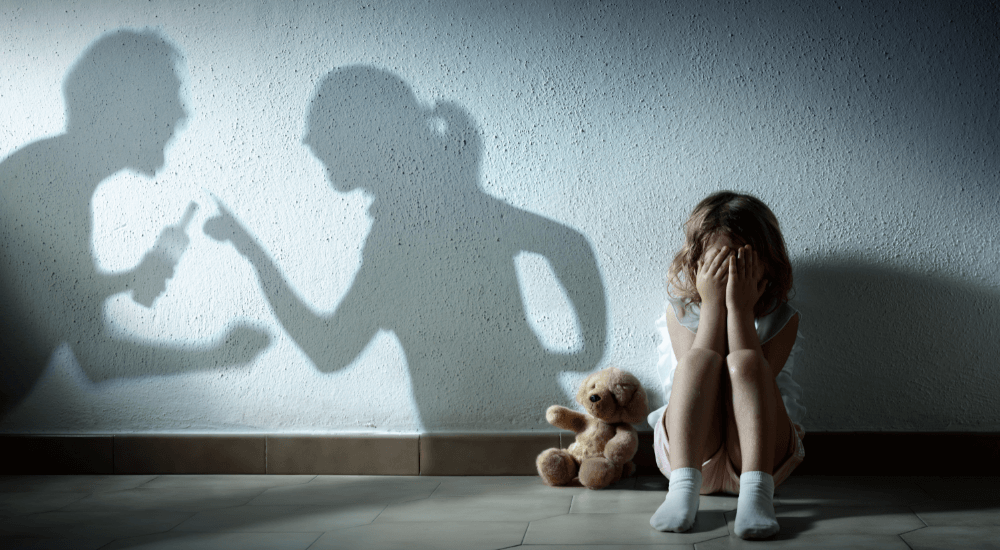 Child covering face, distressed by shadows of arguing adults on wall. Teddy bear nearby. Concept of domestic conflict impact.