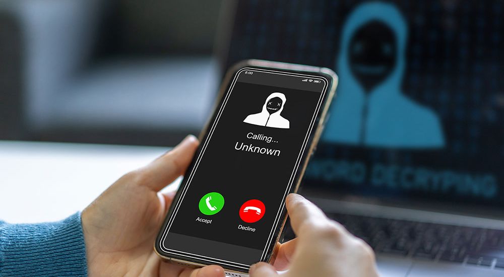 Person holding a phone showing an unknown call, with a hacker image on the laptop in the background.