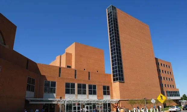 Modern red-brick building with large windows under a clear blue sky, people walking in front, sunny day.