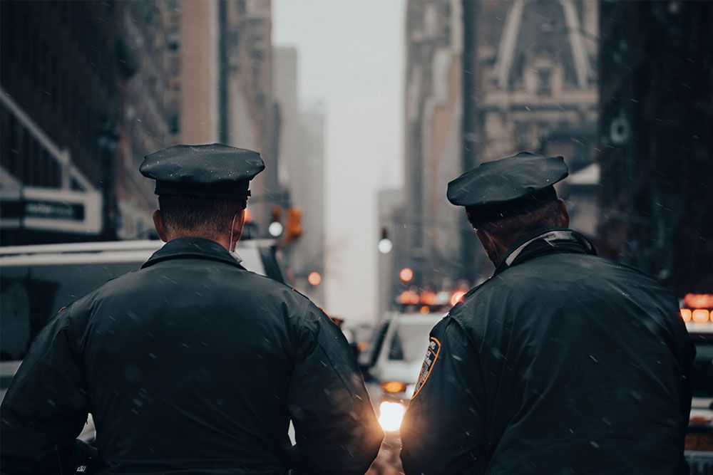 Two police officers patrolling a snowy city street, facing away, with buildings and lights in the background.