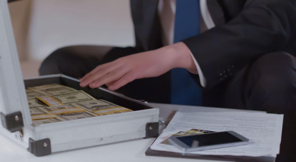 Person in a suit opens briefcase full of cash beside documents and smartphone on table.