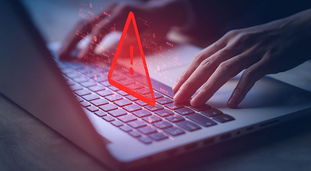 Hands typing on a laptop keyboard with a red warning symbol, indicating cyber threat or computer virus alert.